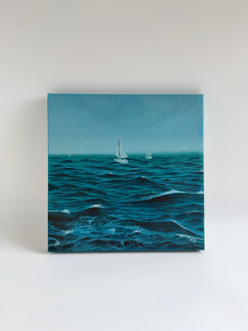 Original stormy sailboat oil painting featuring a lone sailing boat navigating dramatic ocean waves on a 12×12 square canvas by Shreya Chandra.